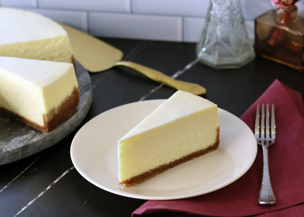 A plain slice of New York Cheesecake on a white plate.