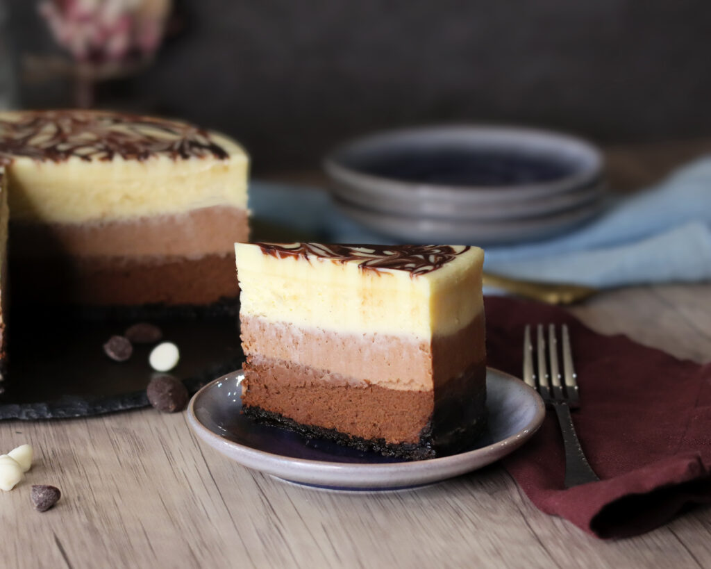 Triple Chocolate Cheesecake slice with the whole cake and stack of plates in the background
