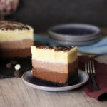 Triple Chocolate Cheesecake slice with the whole cake and stack of plates in the background