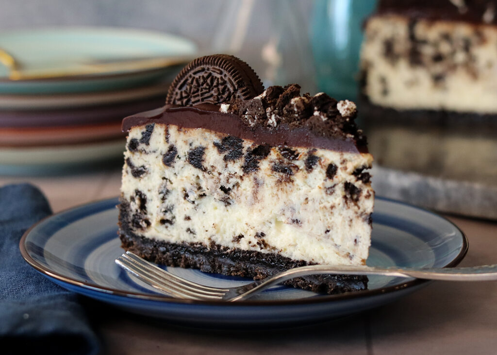 Cookies'N Cream Cheesecake slice on a blue plate in front of a stack of more plates and the whole pie.
