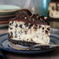 Slice of Cookies ’N Cream cheesecake on a plate, showing the Oreo crust, creamy Oreo‑studded filling, and chocolate ganache topped with an Oreo cookie.