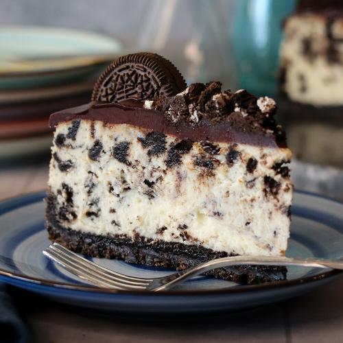 Slice of Cookies ’N Cream cheesecake on a plate, showing the Oreo crust, creamy Oreo‑studded filling, and chocolate ganache topped with an Oreo cookie.