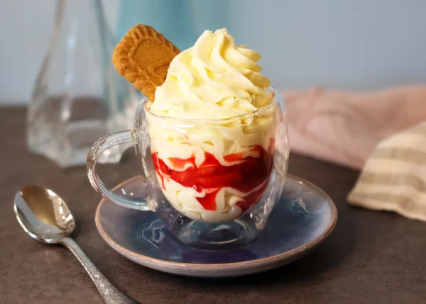 A glass cup of cheesecake mousse with a clearly visible layer of macerated strawberries and a cookie garnish.