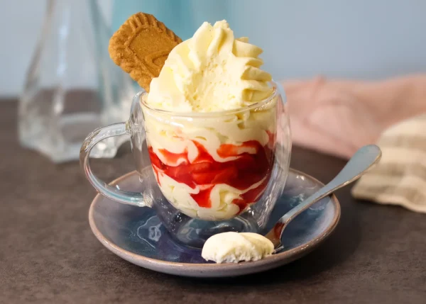 A glass cup of cheesecake mousse with a clearly visible layer of macerated strawberries and a cookie garnish. A missing bite shows the interior texture of the mousse.
